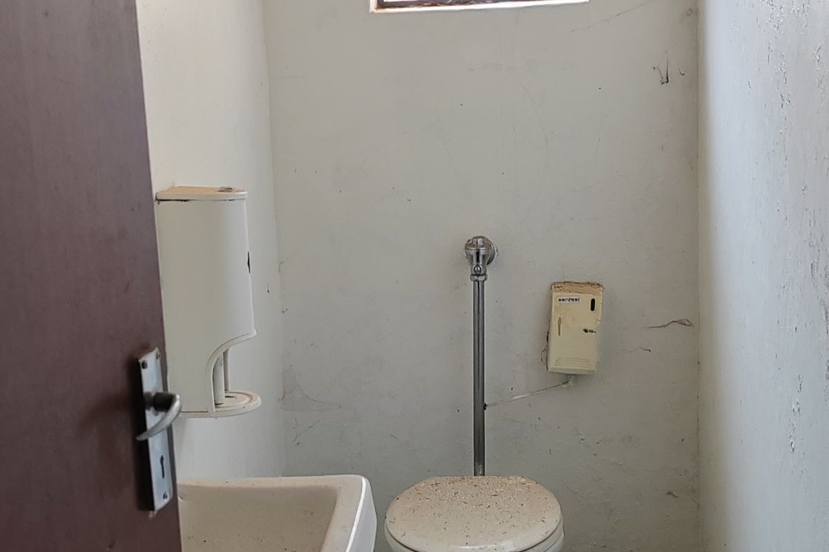 A small industrial-style bathroom featuring a basic toilet, wall-mounted basin, and security-barred window, requiring maintenance and cosmetic updates.