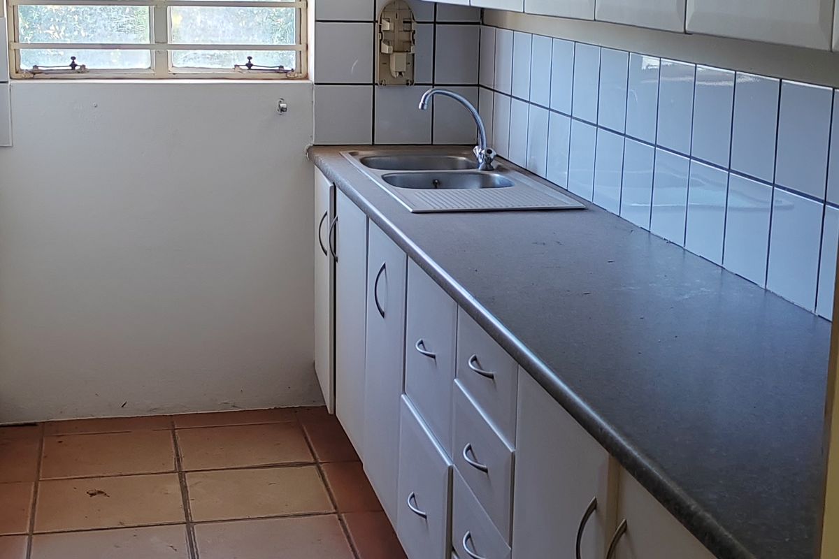 A functional kitchen area featuring built-in cabinetry, a stainless steel sink, tiled splashback, and ceramic floor tiles with security bars on the window.