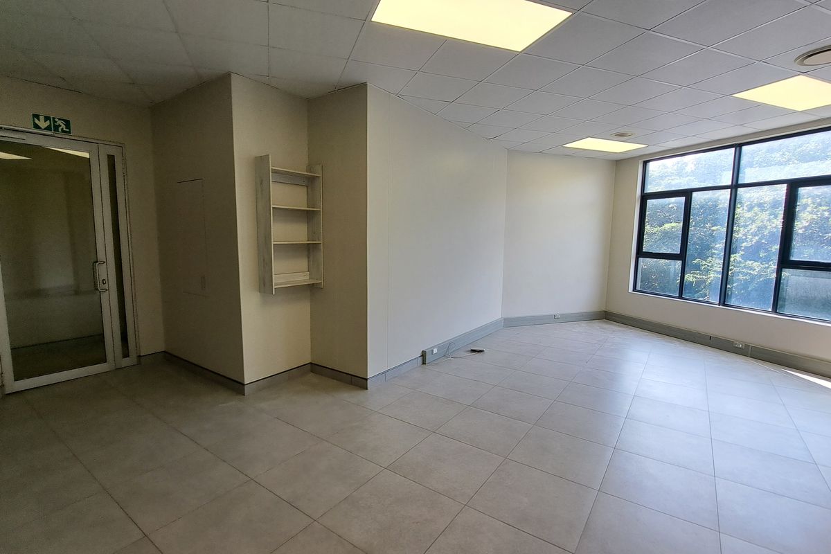A bright, open-plan office space featuring large windows, neutral tiled flooring, and a modern suspended ceiling with integrated lighting.