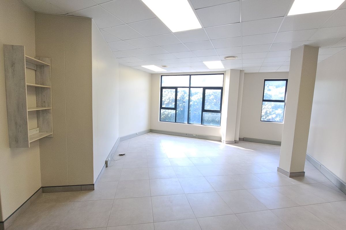 A bright, open-plan office space featuring neutral tiled flooring, a suspended ceiling with recessed lighting, and large windows providing ample natural light.