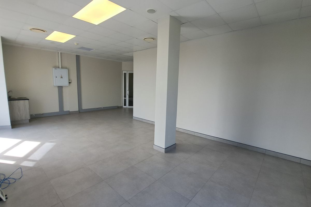 An open-plan office space featuring tiled flooring, a suspended ceiling with recessed lighting, a structural column, and a small built-in kitchenette area.
