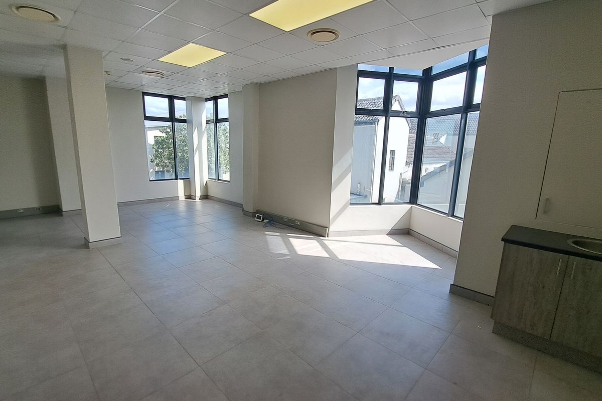 A bright, open-plan office space featuring tiled flooring, a suspended ceiling with recessed lighting, large windows, and a compact built-in kitchenette area.