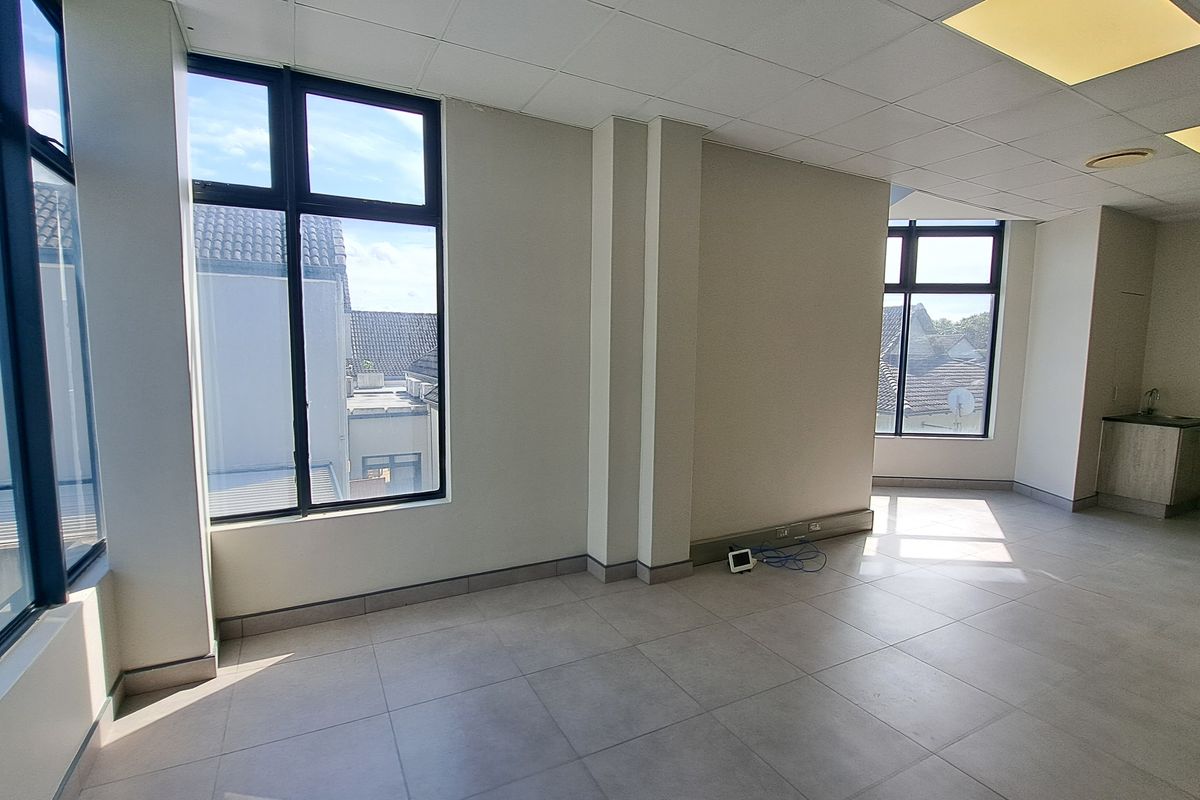 A bright, open-plan office space featuring large windows, tiled flooring, a suspended ceiling with recessed lighting, and a compact kitchenette area.