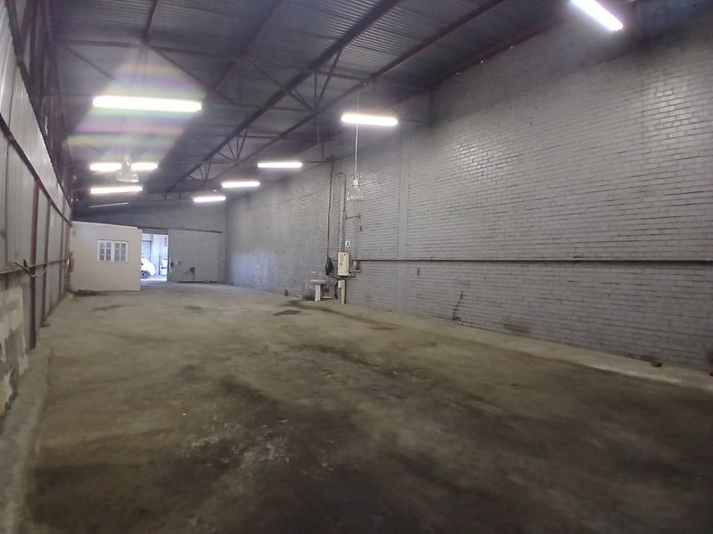 Spacious industrial utility area featuring exposed steel roof trusses, concrete flooring, and wall-mounted lighting, suitable for storage or workshop use.