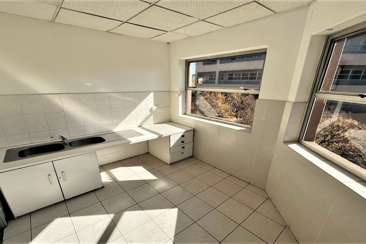 A functional office kitchenette featuring tiled flooring, wall tiling, a built-in sink, and cabinetry with ample natural light from large windows.