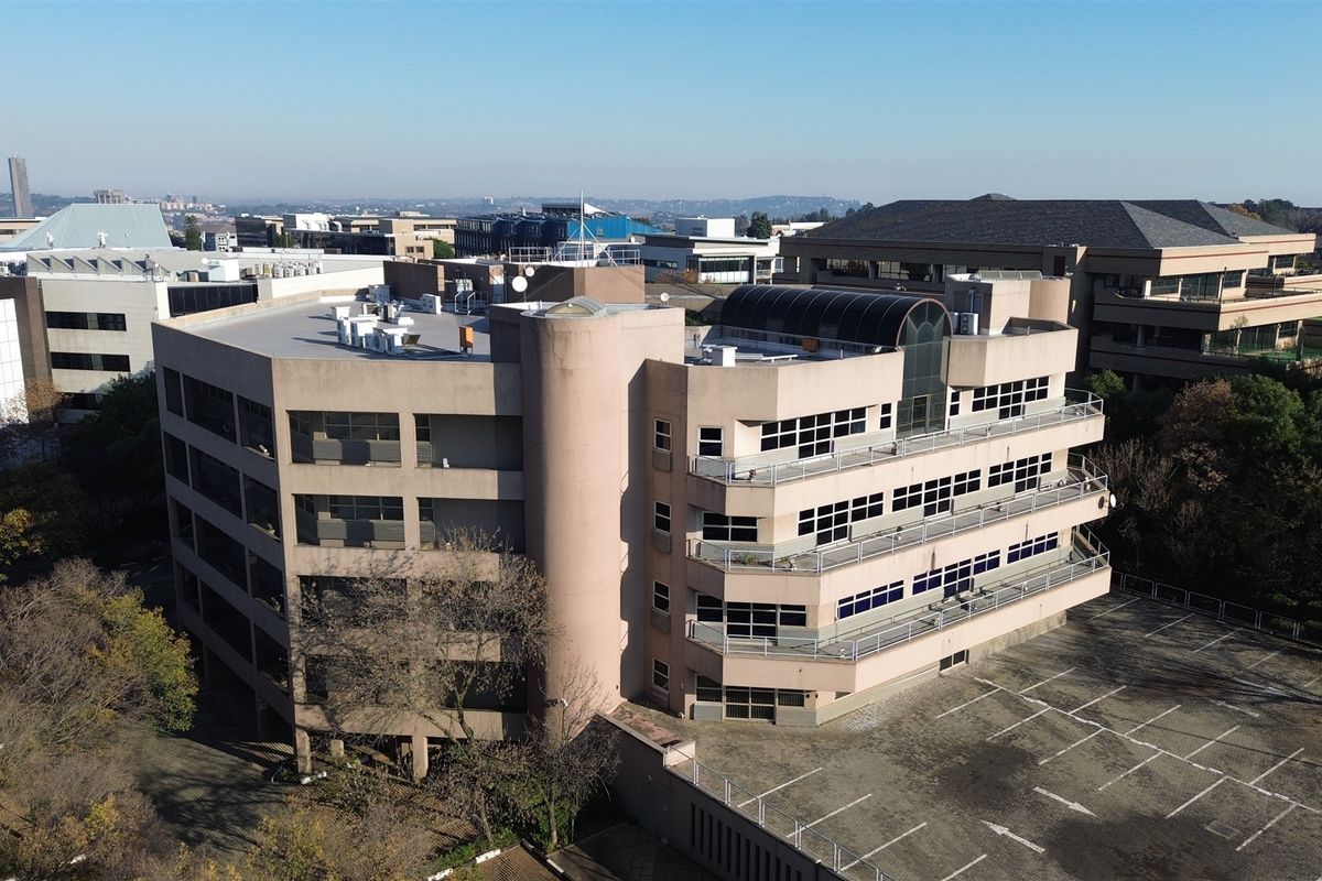 Aerial view of a multi-story commercial office building featuring tiered balconies, flat roof with HVAC units, and an adjacent paved parking lot.