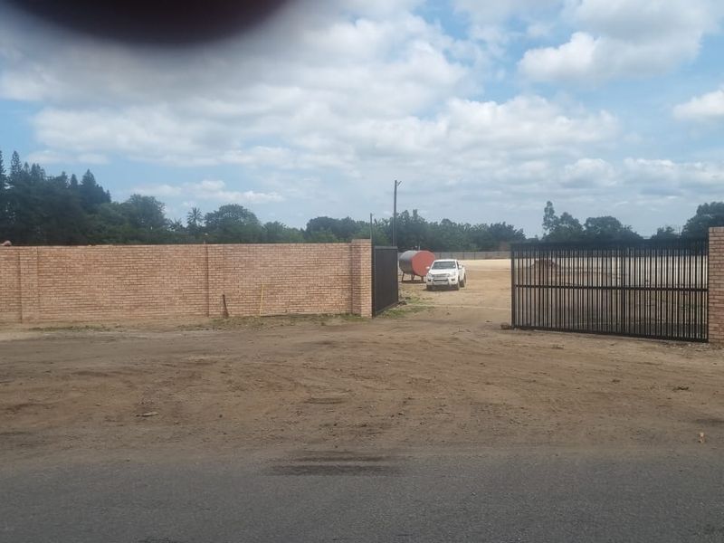 A view of the industrial property entrance featuring a brick perimeter wall and a large metal security gate leading to an unpaved yard.
