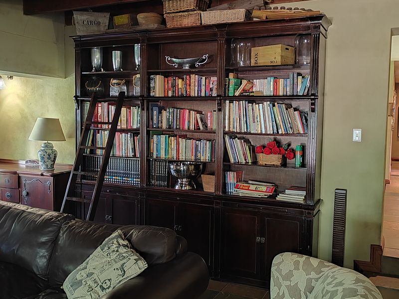 A cozy living room featuring a large built-in wooden library unit with a sliding ladder, exposed timber ceiling beams, and durable stone tile flooring.