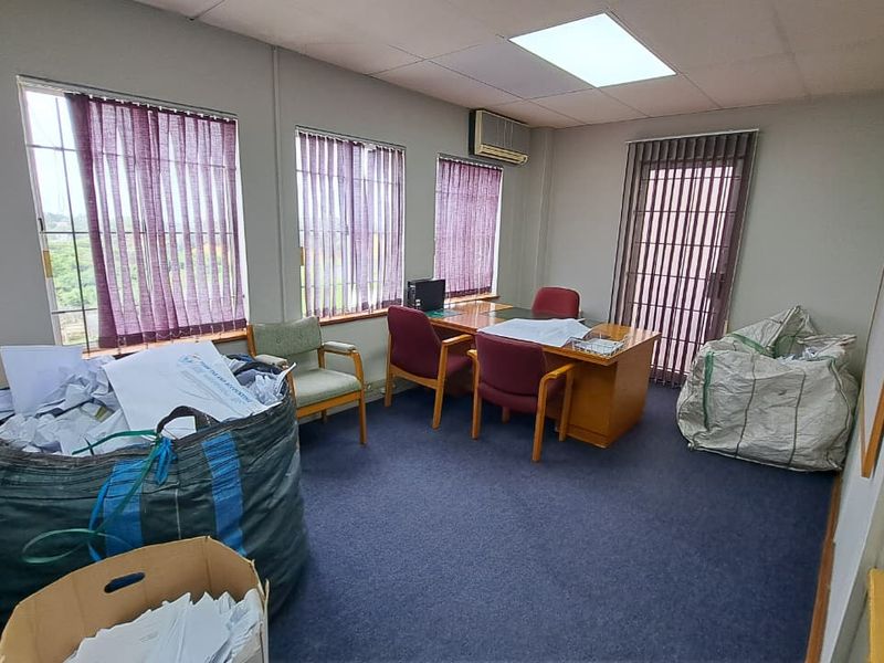 A functional office space featuring carpet flooring, drop ceiling with recessed lighting, wall-mounted air conditioning, and large windows with vertical blinds.