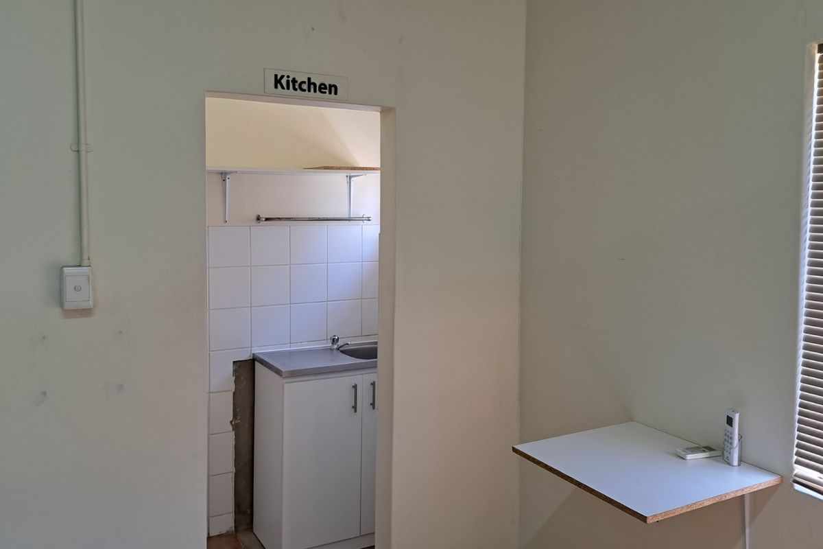 A small office kitchenette featuring a single sink, base cabinetry, and tiled splashback, adjacent to a wall-mounted desk area.