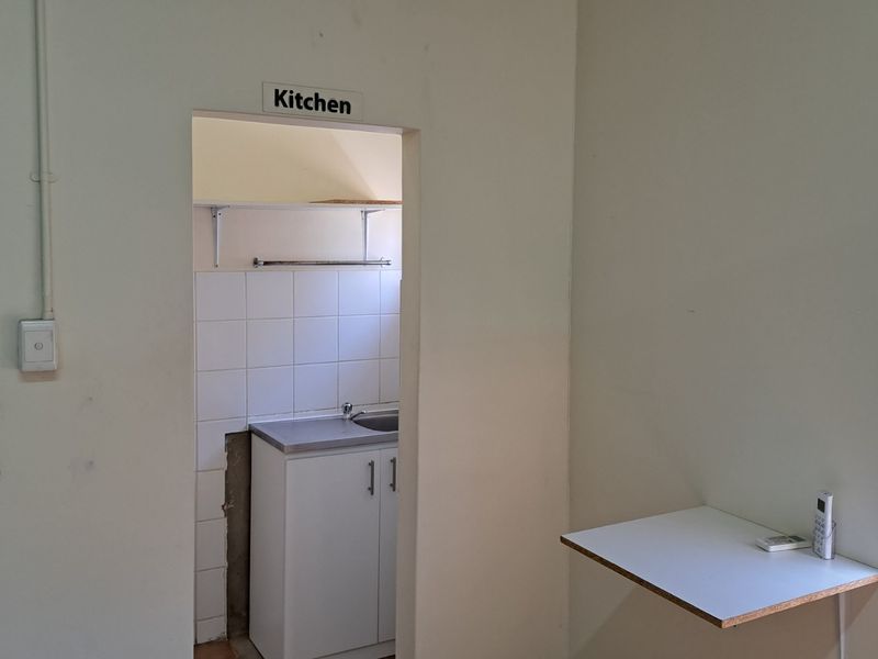 A small office kitchenette featuring a single sink, base cabinetry, and tiled splashback, adjacent to a wall-mounted desk area.