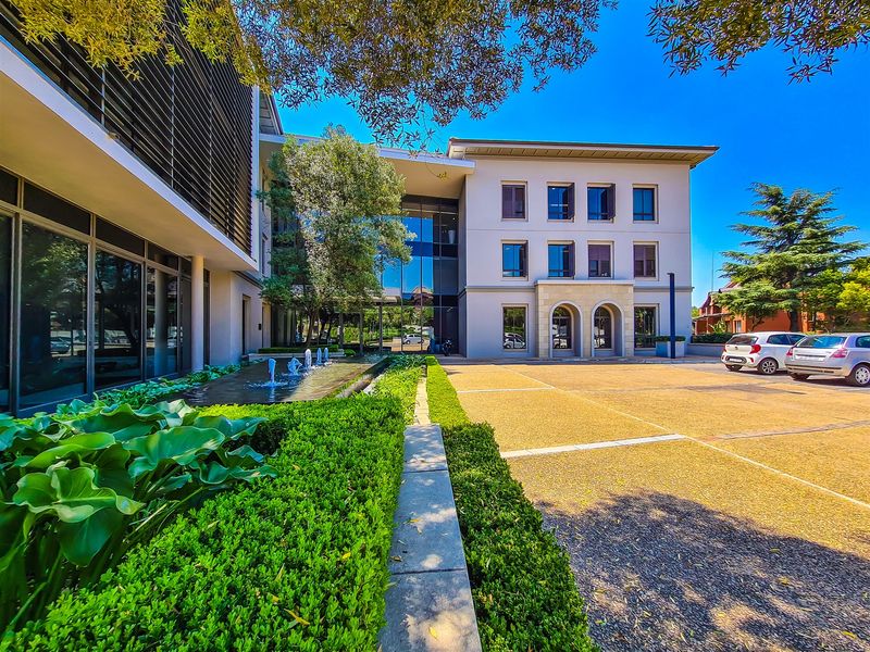 Modern office building exterior featuring a landscaped garden, water feature, paved parking area, and contemporary architectural design with glass and stone elements.