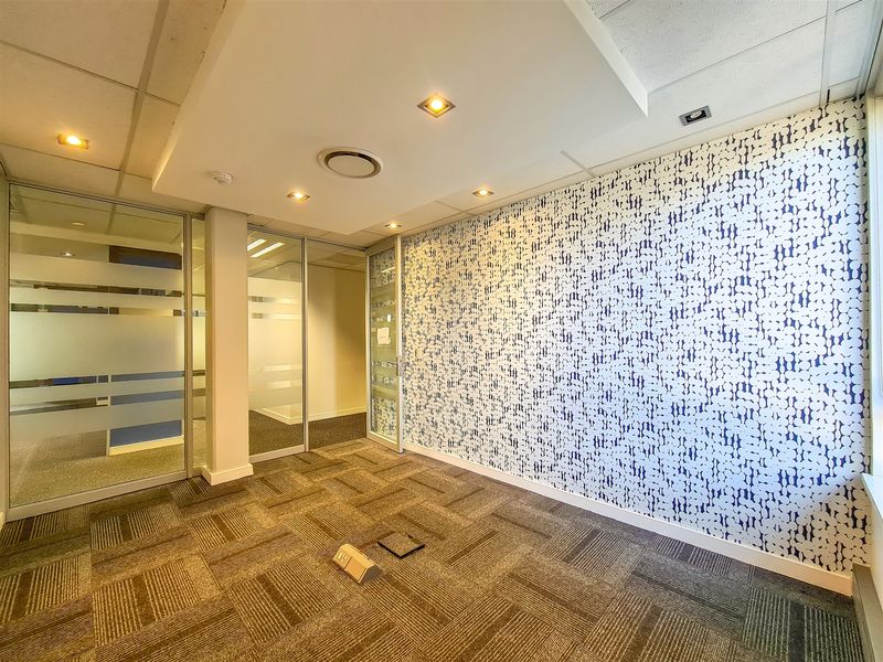 A bright, empty office space featuring modern glass partitions, patterned feature wall, recessed ceiling lighting, and durable carpet tiles.