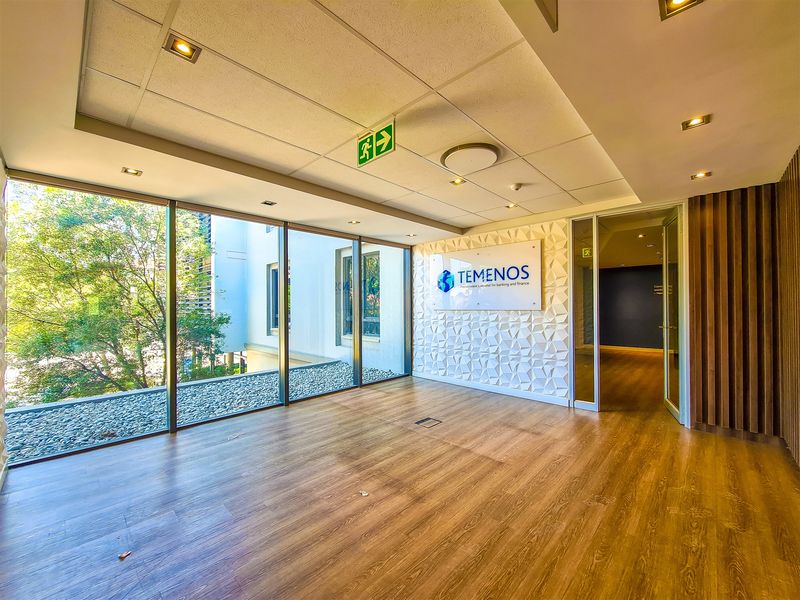 A bright, modern office reception area featuring floor-to-ceiling windows, wood-look flooring, a textured feature wall with branding, and recessed ceiling lighting.