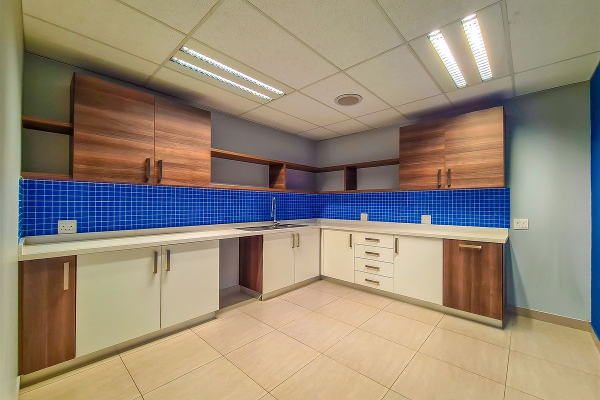 A clean, modern office kitchenette featuring two-tone cabinetry, blue tiled backsplash, stainless steel sink, and durable ceramic floor tiles.