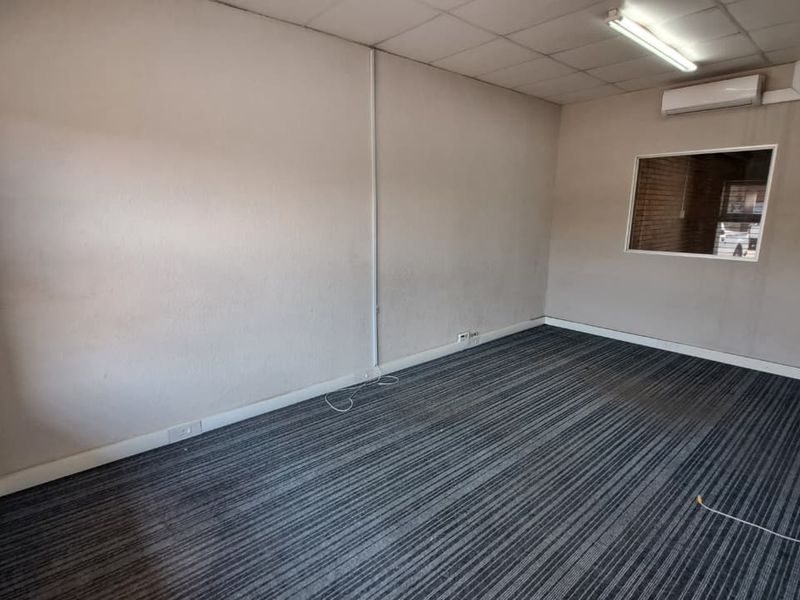 An empty office space featuring carpeted flooring, a suspended ceiling with fluorescent lighting, a wall-mounted air conditioning unit, and an internal window.