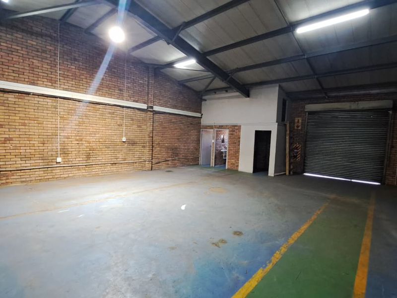 Spacious industrial warehouse unit featuring exposed brick walls, concrete flooring, high ceilings with steel trusses, and a large roller shutter door for easy access.