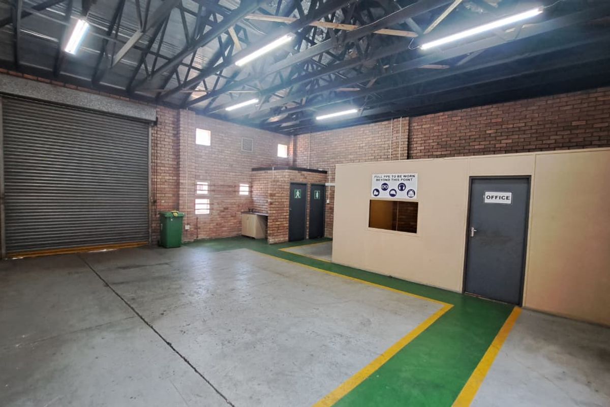 Spacious industrial utility area featuring exposed steel roof trusses, facebrick walls, concrete flooring, a large roller shutter door, and a partitioned office space.