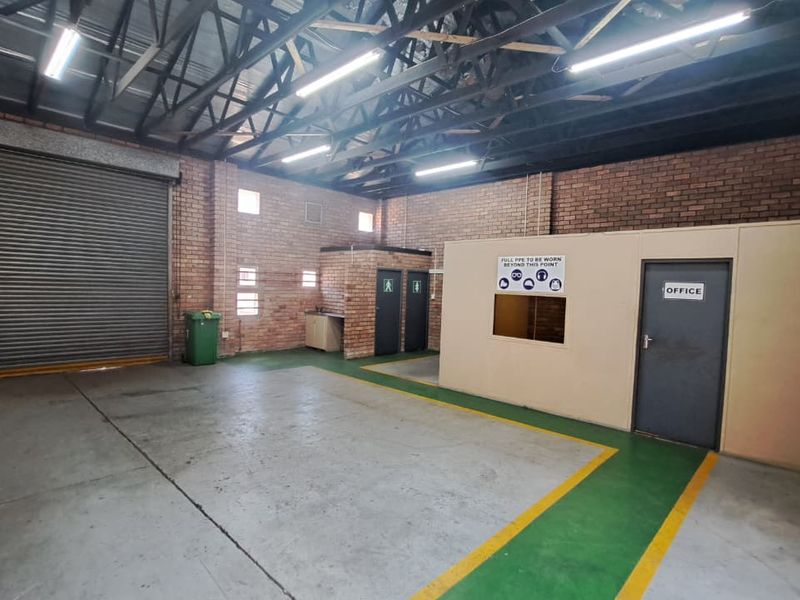 Spacious industrial utility area featuring exposed steel roof trusses, facebrick walls, concrete flooring, a large roller shutter door, and a partitioned office space.