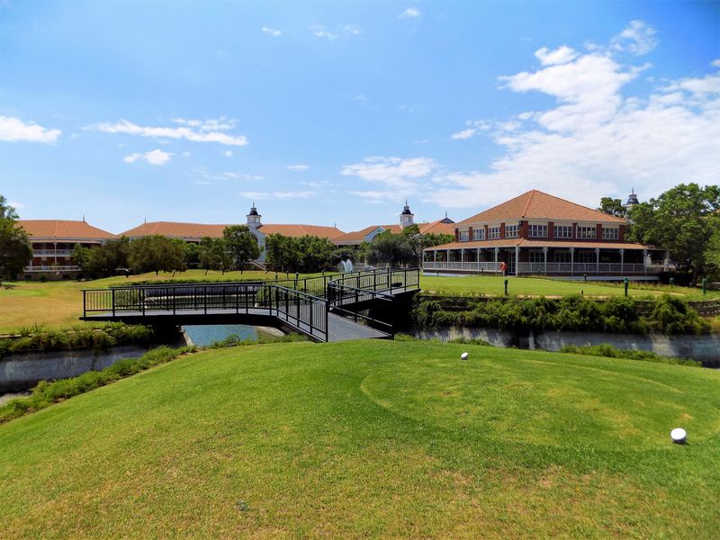 A scenic view of the office property grounds featuring a well-maintained lawn, a pedestrian bridge over a water feature, and a large building with a veranda.