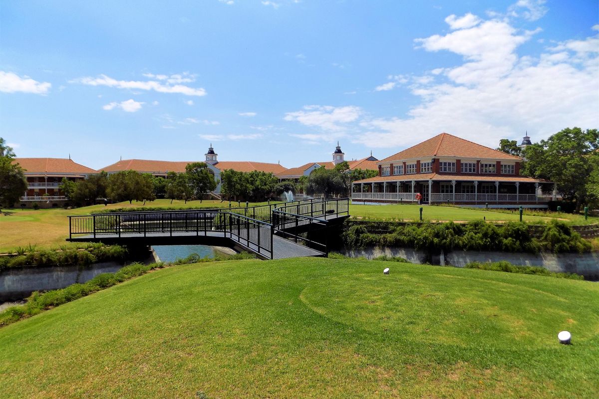 A scenic view of the office property grounds featuring a well-maintained lawn, a pedestrian bridge over a water feature, and a large building with a veranda.