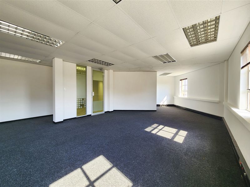 A spacious, empty office area featuring carpeted flooring, a suspended ceiling with integrated lighting, and a glass-paneled door.
