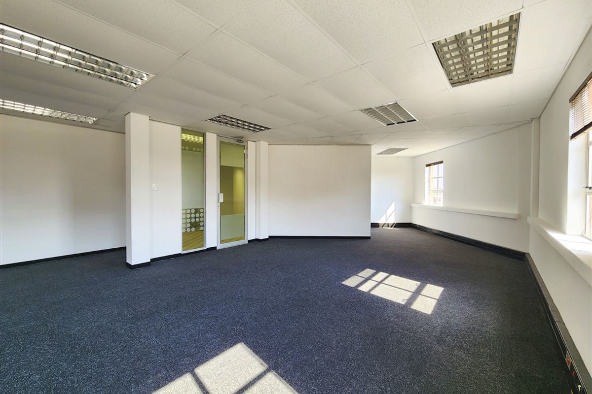 A spacious, empty office area featuring carpeted flooring, a suspended ceiling with integrated lighting, and a glass-paneled door.