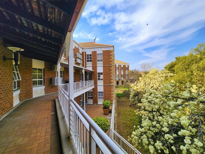A spacious covered balcony overlooking a landscaped garden area, featuring brick exterior walls and secure metal railings in a well-maintained office complex.