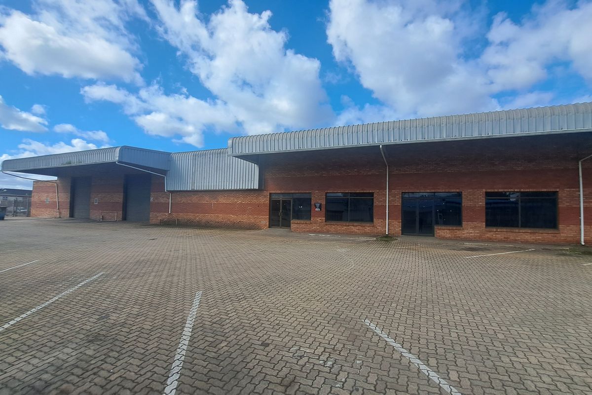 Industrial property featuring a brick facade, metal roofing, multiple roller shutter doors, and an expansive paved parking area for logistics or commercial use.
