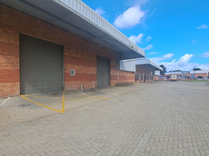 Industrial warehouse exterior featuring multiple roller shutter doors, brick facade, and a large paved loading area under a metal canopy.