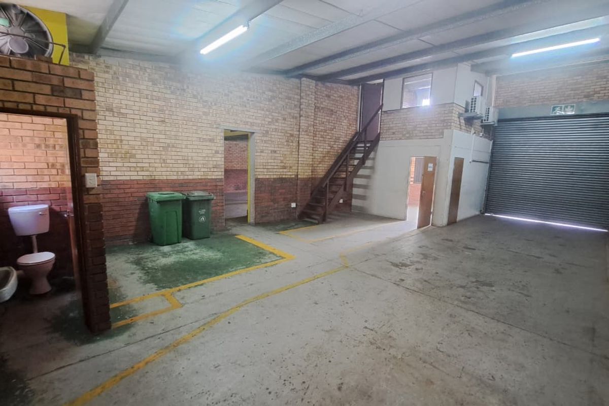 Industrial warehouse space featuring exposed brick walls, concrete flooring, a roller shutter door, mezzanine access, and a basic integrated toilet facility.