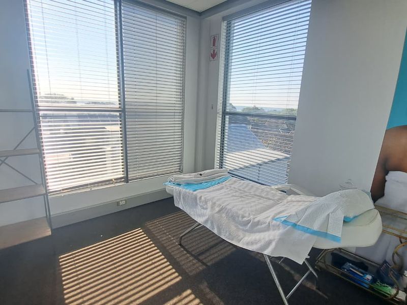 A bright office or treatment room featuring large windows with venetian blinds and carpeted flooring, suitable for professional use.