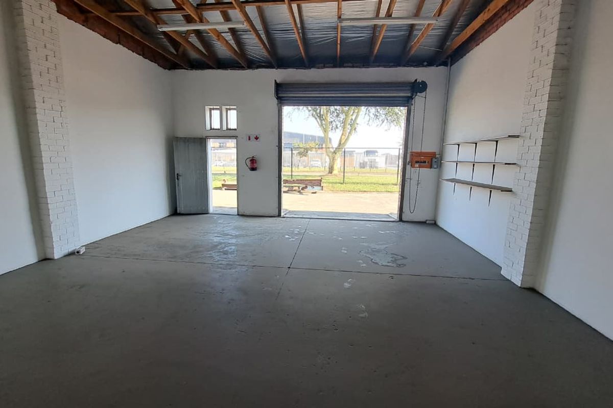 Spacious industrial utility space featuring concrete flooring, exposed timber roof trusses, a large roller shutter door, and wall-mounted shelving.
