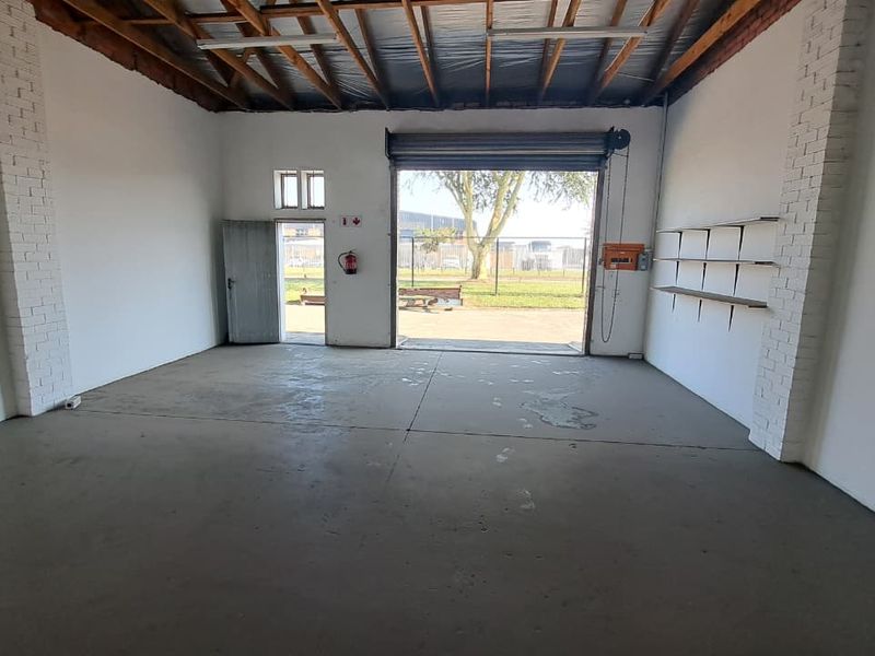 Spacious industrial utility space featuring concrete flooring, exposed timber roof trusses, a large roller shutter door, and wall-mounted shelving.
