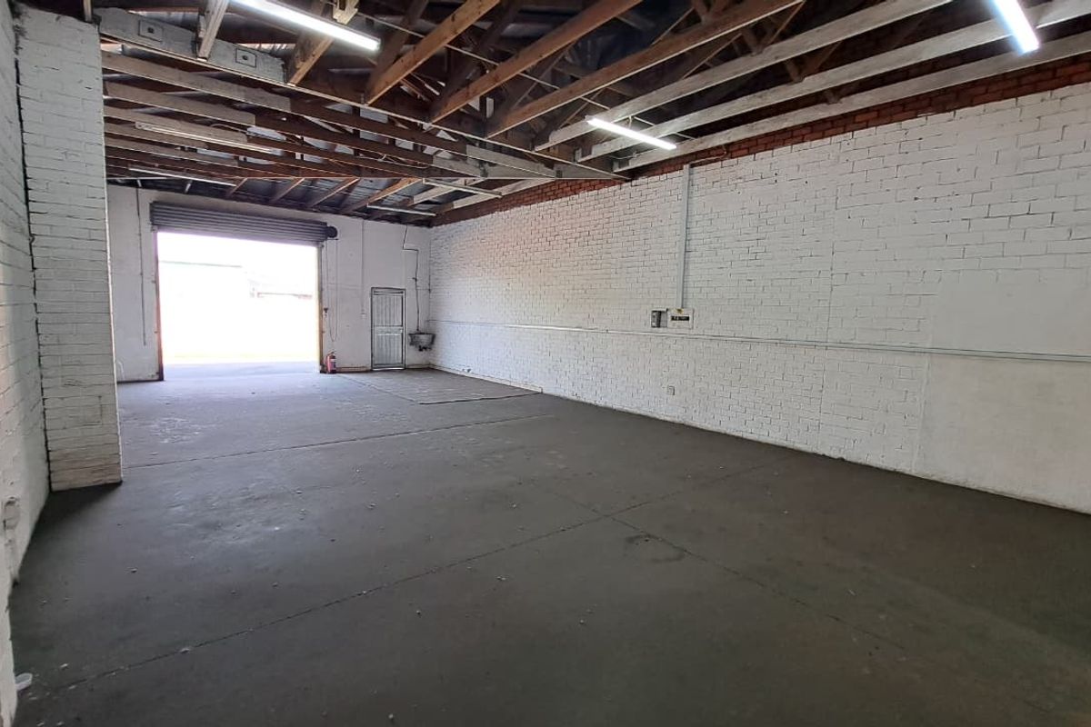 Spacious industrial garage featuring exposed timber roof trusses, painted brick walls, concrete flooring, and a large roller shutter door for vehicle or equipment access.