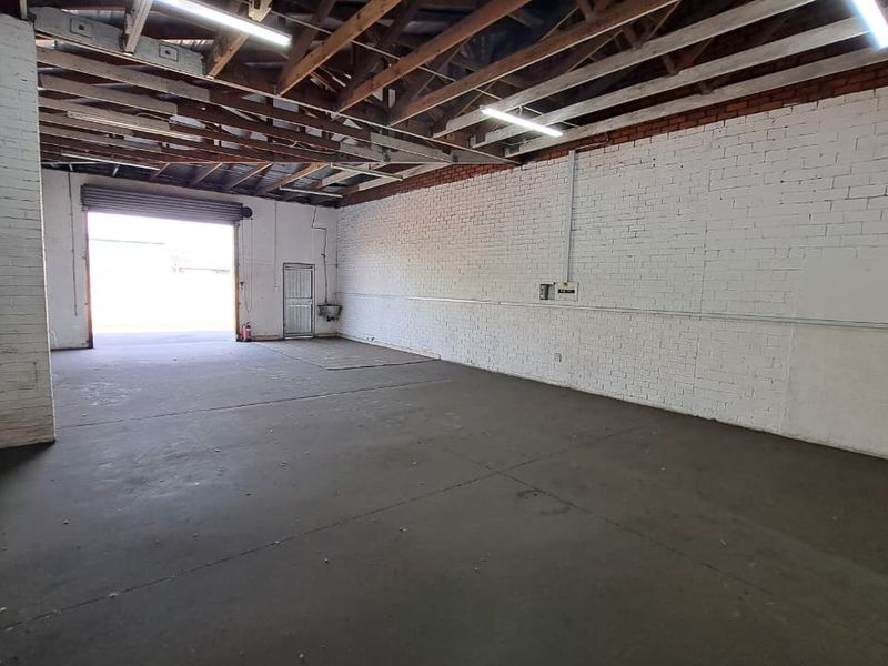 Spacious industrial garage featuring exposed timber roof trusses, painted brick walls, concrete flooring, and a large roller shutter door for vehicle or equipment access.