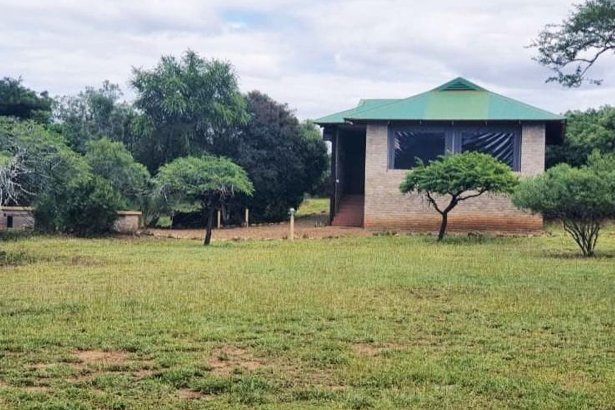 A view of a brick-built farm cottage featuring a green metal roof and a covered patio, set within a spacious, natural garden landscape.