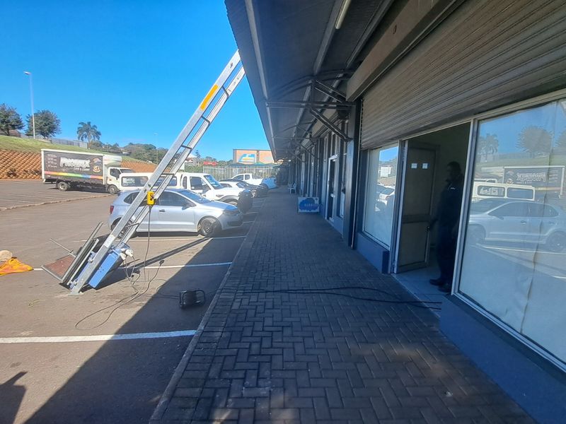 Exterior view of a retail storefront featuring a paved walkway, glass frontage, and roller shutter doors with adjacent parking facilities.