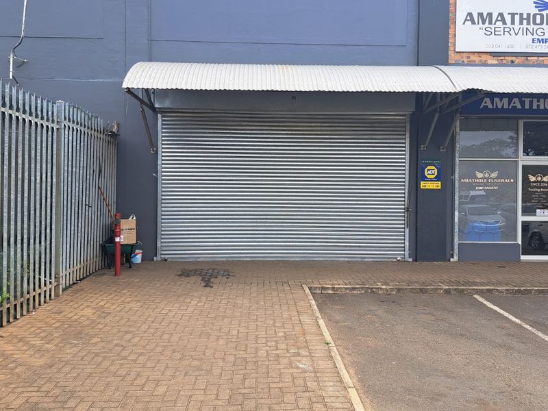 Front exterior view of a commercial retail unit featuring a secure roller shutter door, paved entrance area, and glass storefront signage.