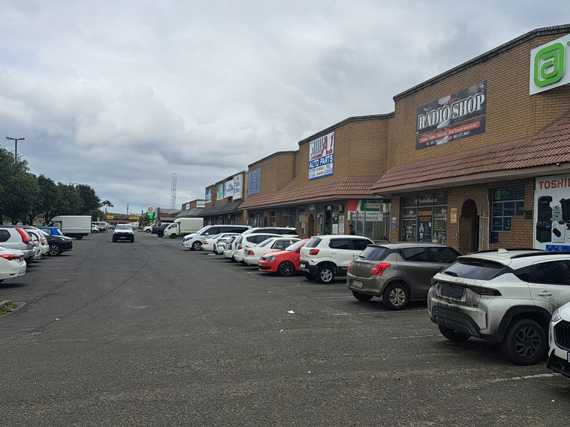 A commercial strip mall featuring multiple storefronts with brick facades, tiled roofing, and an extensive paved parking area for customers.