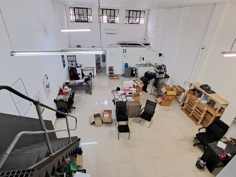 Open-plan commercial space featuring tiled flooring, high-level windows, a metal staircase, and a small kitchenette area, suitable for office or retail use.