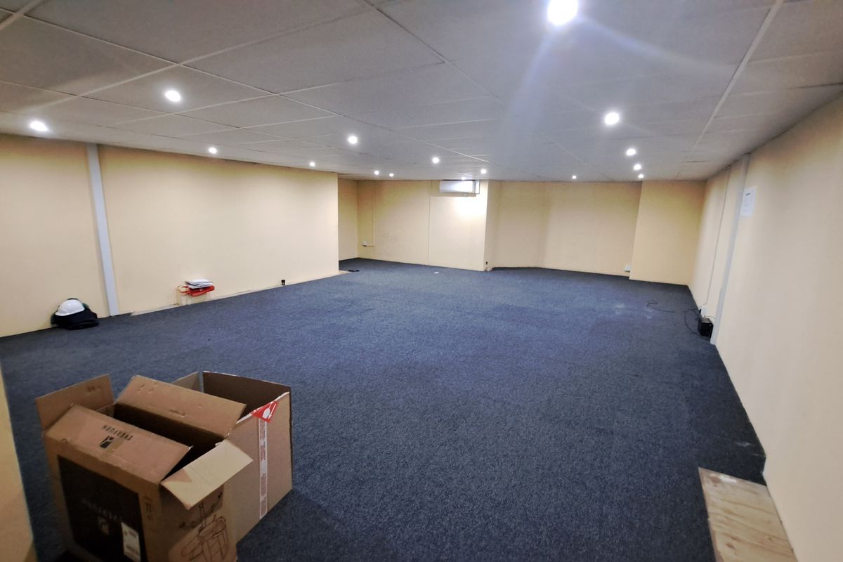 A spacious, empty commercial office area featuring a suspended ceiling with recessed lighting, wall-mounted air conditioning, and dark carpet flooring.