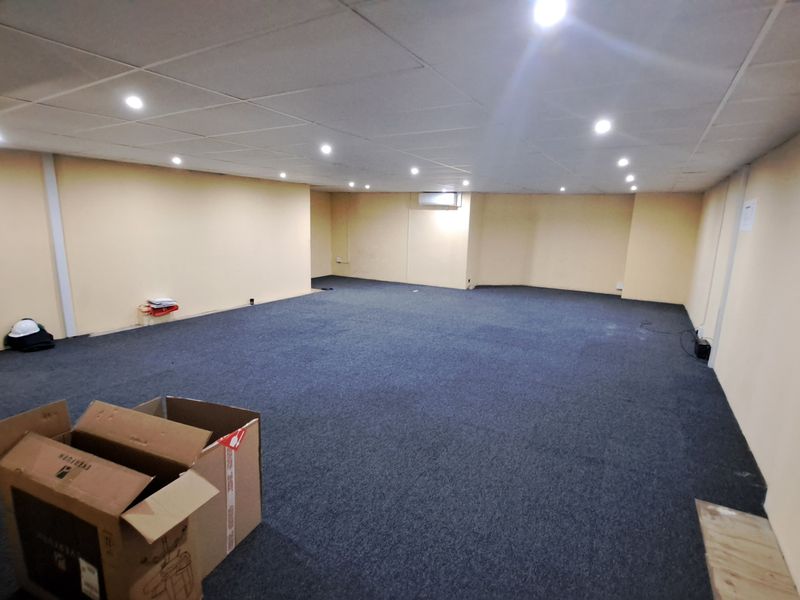 A spacious, empty commercial office area featuring a suspended ceiling with recessed lighting, wall-mounted air conditioning, and dark carpet flooring.