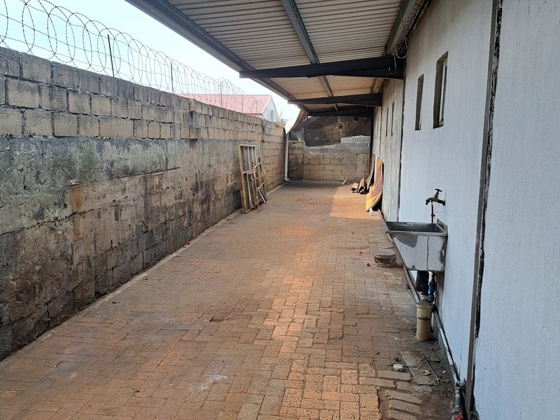 A narrow exterior rear service area featuring a paved walkway, a wall-mounted utility sink, and a protective metal canopy along the building side.