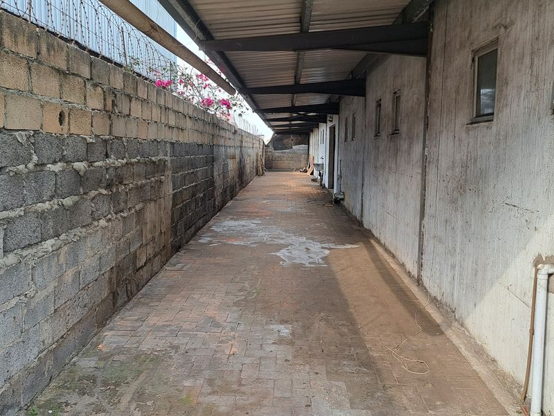 A narrow, paved exterior side passage featuring a covered walkway, cinder block boundary wall with security fencing, and weathered industrial wall finishes.