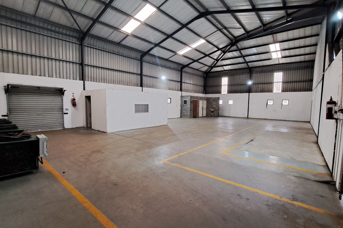 Spacious industrial warehouse featuring high ceilings with exposed steel trusses, concrete flooring, roller shutter access, and integrated partitioned office or utility space.