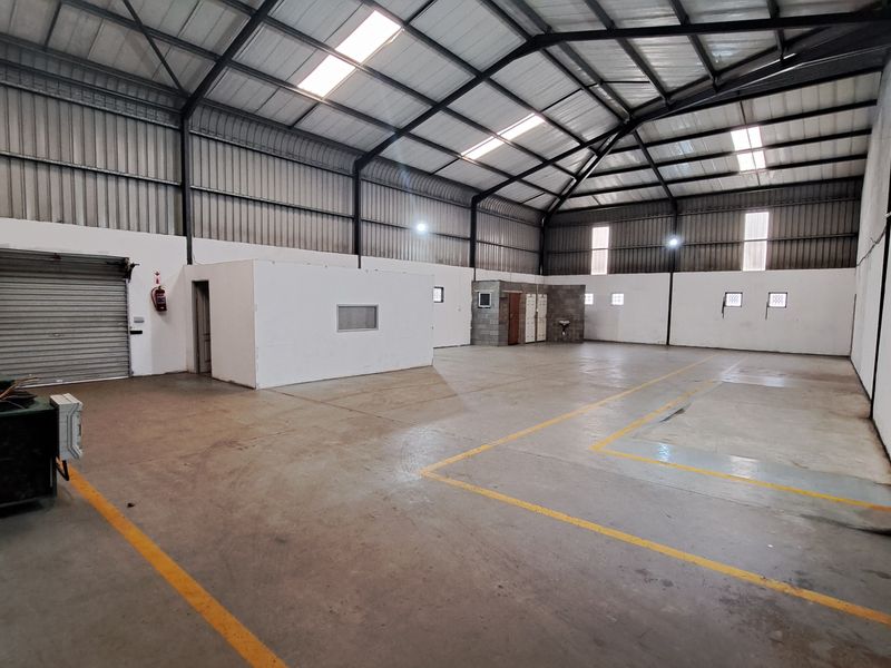 Spacious industrial warehouse featuring high ceilings with exposed steel trusses, concrete flooring, roller shutter access, and integrated partitioned office or utility space.