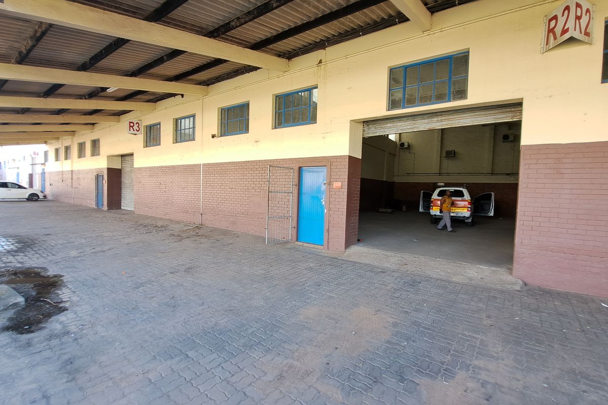 Industrial property exterior featuring a covered loading bay, paved driveway, and multiple roller shutter access points for logistics or warehouse operations.
