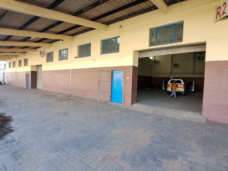 Industrial property exterior featuring a covered loading bay, paved driveway, and multiple roller shutter access points for logistics or warehouse operations.