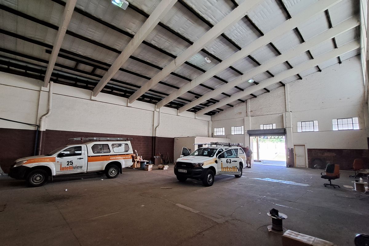 Spacious industrial warehouse or garage area featuring high ceilings with exposed steel trusses, concrete flooring, and large roller shutter access.